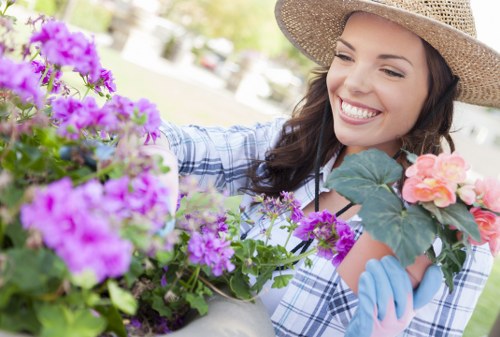 A woman wearing a straw hat and blue gardening gloves is gardening in an outdoor space, surrounded by vibrant pink, purple, and peach flowering plants in a garden with a well-maintained grassy lawn. In the background, there are additional garden elements such as trees and shrubs, with sunlight filtering through, indicating a bright and clear day. The garden features a landscaped layout with flower beds, a paved area, and a well-kept lawn, which highlights outdoor care and landscaping efforts typical of gardens in Shepherds Bush. This scene reflects routine gardening and maintenance activities carried out by Landscapers Shepherds Bush, emphasizing the natural tones, diverse plant textures, and organized outdoor environment in a typical residential garden setting in London postcode W12, supporting their gardening services.