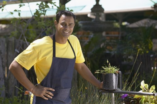 A smiling man wearing a yellow t-shirt and a dark blue apron stands in a well-maintained garden, holding a small black plastic pot with a young plant. Behind him, there is a lush green hedge, flowering plants with purple and yellow blossoms, and a wooden fence that encloses the garden area. To the right, a raised outdoor table holds a gardening tray or container, while in the background, a pergola or garden structure is partially visible, shaded by a green canopy. The garden features a mix of grass lawn and paved pathways, with natural light indicating a bright, clear day. This scene exemplifies outdoor maintenance and gardening activities often supported by professional landscapers in Shepherds Bush, contributing to tidy and vibrant backyard spaces.