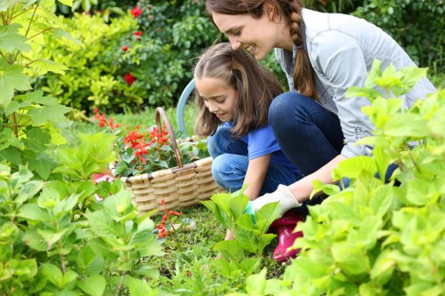 A woman and a young girl are gardening together in a lush, well-maintained outdoor space, likely part of a residential garden in Shepherd's Bush. They are kneeling on the grass amidst vibrant green foliage and flowering plants, with the girl holding a small gardening basket filled with red blooms. The garden features a combination of dense shrubs, leafy plants, and a neatly trimmed lawn area with a smooth surface, surrounded by natural and cultivated plant beds. In the background, there are taller bushes and trees providing shade and a sense of enclosure. The sunlight is soft, indicating a partly cloudy day, enhancing the natural colours of the greenery and flowers. The scene reflects outdoor maintenance and gardening care, potentially carried out by professionals such as those from Landscapers Shepherds Bush, contributing to the tidy, inviting appearance of the landscaped yard.