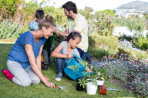 A family gardening outdoors in a lush, well-maintained garden with a variety of flowering plants, shrubs, and small trees. In the foreground, a woman with a fork is tending to the grass, while a young girl is watering plants from a blue container. Nearby, gardening tools and pots are arranged on the lawn, which features neatly trimmed grass and flower beds bordered with stones. Behind them, a man and a girl are sitting on a low garden wall, surrounded by dense foliage and flowering plants, with a view of distant hills and a partly cloudy sky in the background. The scene captures a sunny day with natural lighting, highlighting the vibrant green tones of the lawn and the colorful blooms, as part of a landscaped outdoor space typical of residential gardens in Shepherds Bush, London, where professional garden maintenance and landscaping services are commonly provided by companies such as Landscapers Shepherds Bush.