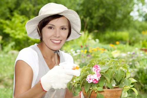 A woman wearing a wide-brimmed white hat and gardening gloves is outdoors in a lush, green garden during daytime, tending to a vibrant pink flowering plant in a terracotta pot. In the background, there are well-maintained grassy lawn areas, flower beds with various colorful blooms, and a backdrop of dense trees and shrubs, suggesting a landscaped outdoor space typical of a private garden in Shepherds Bush. The scene is well-lit with natural sunlight, highlighting the healthy, green foliage and bright flowers, while the woman appears cheerful and engaged in gardening activities, reflecting professional gardening and landscaping services offered by Landscapers Shepherds Bush in the local area.