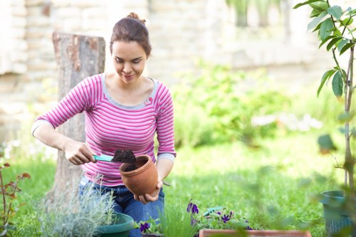A woman with dark hair tied back, wearing a pink and white striped long-sleeve shirt, is working in a garden. She is holding a terracotta plant pot with soil inside and using a small trowel to add soil to the pot. The garden features a lush green lawn in the foreground, bordered by a variety of potted plants and flowers, including purple blooms. In the background, a wooden fence runs along the garden perimeter, with a tree trunk visible on the left and some bushes on the right. The outdoor space appears well-maintained with natural daylight illuminating the scene, suggesting a pleasant weather day. This setting is typical of a suburban garden in Shepherds Bush, and reflects gardening activities undertaken by professional landscapers like Landscapers Shepherds Bush, including planting and garden maintenance, which supports their expertise in gardening services for the local area.