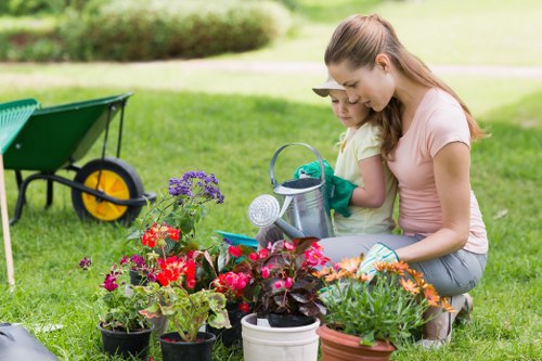 A woman and a young girl are kneeling together on a lush green lawn in a garden, engaging in planting flowers. The garden features a well-maintained grassy area with a variety of colorful flowering plants in pots, including shades of red, pink, purple, and orange. In the background, there is a paved area or patio and a wooden wheelbarrow with yellow wheels, positioned nearby for gardening tasks. The scene is set outdoors on a bright, clear day with natural sunlight illuminating the space, which appears to be a back garden or outdoor landscaped area typical of residential properties in Shepherds Bush. The garden elements include flower beds, potted plants, and a tidy lawn, highlighting the outdoor maintenance and gardening services offered by Landscapers Shepherds Bush in the local area, with a focus on nurturing and beautifying outdoor spaces.