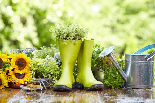 A pair of bright green rubber gardening boots filled with small white flowering plants are positioned on a wooden garden table, with a background of lush green foliage indicating an outdoor garden setting. To the left, there is a colorful arrangement of sunflowers and other flowering plants, adding vibrant yellow and purple accents. To the right, a metal watering can with a blue handle rests on the table beside the boots, and a small garden trowel with a tarnished blade lies on the surface. The scene suggests a well-maintained garden space suitable for activities like planting, watering, and general outdoor gardening. The natural light highlights the textures of the wooden table and the various plants and tools, emphasizing the outdoor environment characteristic of professional gardening and landscaping services such as those provided in Shepherds Bush. This detailed and naturalistic description aligns with a gardening service webpage focused on sustainable practices and garden maintenance, supported by the local context of West London.