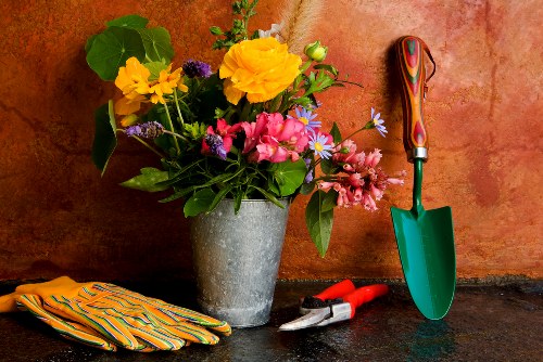 A still life arrangement on a dark surface featuring a large, grey stone-effect pot filled with a vibrant mix of garden flowers, including yellow, pink, purple, and white blooms, with green foliage. To the right of the pot, a small green hand trowel with a wooden handle is positioned upright, alongside a pair of red-handled pruning shears resting on the surface. On the left, a folded pair of multicoloured gardening gloves with yellow, orange, and green stripes are laid out. The background is a textured, warm terracotta wall, creating a natural, earthy setting that reflects outdoor gardening activities and tools used in cultivating flower beds or borders typical of gardens in Shepherd's Bush and surrounding areas.