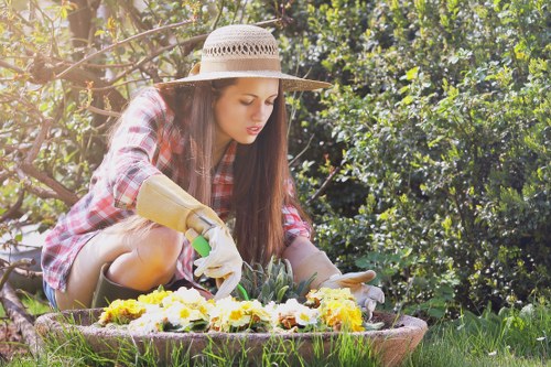 A young woman wearing a wide-brimmed straw hat, a plaid shirt, and gardening gloves is kneeling on a lush green lawn in a backyard garden. She is carefully planting or tending to yellow and white flowering plants within a shallow, circular flower bed bordered by natural soil. Surrounding her, there are dense leafy bushes and trees that provide shade and privacy, creating a vibrant and natural outdoor environment. The garden area features well-maintained grass, and the scene suggests an early morning or late afternoon setting with soft natural sunlight filtering through the foliage. This image highlights outdoor gardening activities typical of residential landscaping and planting services in Shepherds Bush, illustrating a well-kept backyard designed for both aesthetic appeal and leisure, aligning with professional gardening and landscape maintenance practices by Landscapers Shepherds Bush.