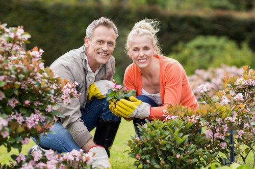 A middle-aged man and woman are seen in a well-maintained garden with lush green grass, flowering shrubs, and pink blossom bushes in the foreground. They are crouched near the plants, smiling at the camera, and appear to be engaged in gardening activities such as pruning or planting. The man is dressed casually in a beige shirt and jeans, while the woman wears a coral top and gardening gloves, suggesting outdoor maintenance work. Behind them, the garden features a neatly trimmed hedge and a variety of trees, with a softly blurred background indicating a sunny day with natural light. The outdoor space is tidy, illustrating a well-kept landscaped yard suitable for gardening services by Landscapers Shepherds Bush, situated within a typical London borough area. This scene emphasizes active garden care, including flowering shrubs and lawn landscaping, reflecting a professional approach to outdoor maintenance and gardening enhancement in Shepherds Bush or nearby West London areas.